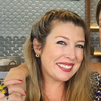 A woman smiling indoors with long blonde hair, wearing red lipstick and a patterned top.